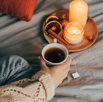 person holding tea in bed with candles, cinnamon sticks and dried oranges